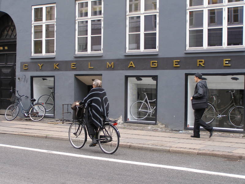 Der Fahrradmacher mit modischer Fahrradfahrerin im Vordergrund.
